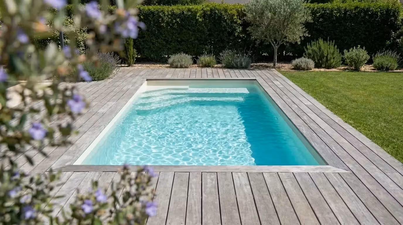 Piscine coque rectangulaire Star 4 entourée d'un deck en bois clair, jardin avec haies taillées et olivier